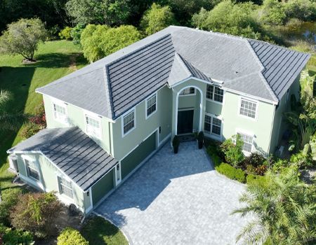 Two-story pale green house with gray roof, three-car garage, and cobblestone driveway surrounded by greenery.
