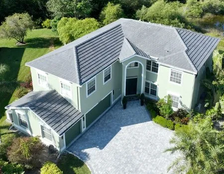 Green two-story house with gray roof and driveway, surrounded by greenery.