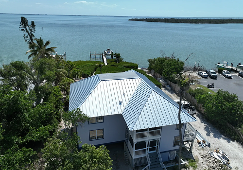 Two-story house with metal roof on waterfront. Dock and boats visible. Overcast sky.