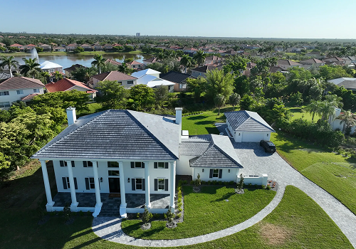 Large white house with columns, gray roof, and long driveway; green lawn and trees surround it.