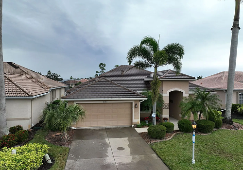 Single-story house with tan stucco exterior, tile roof, and attached garage; palm trees and green lawn in front.