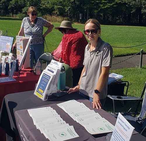 People at an outdoor information table. Woman behind table with brochures, two other people browsing in the background. Sunny day.