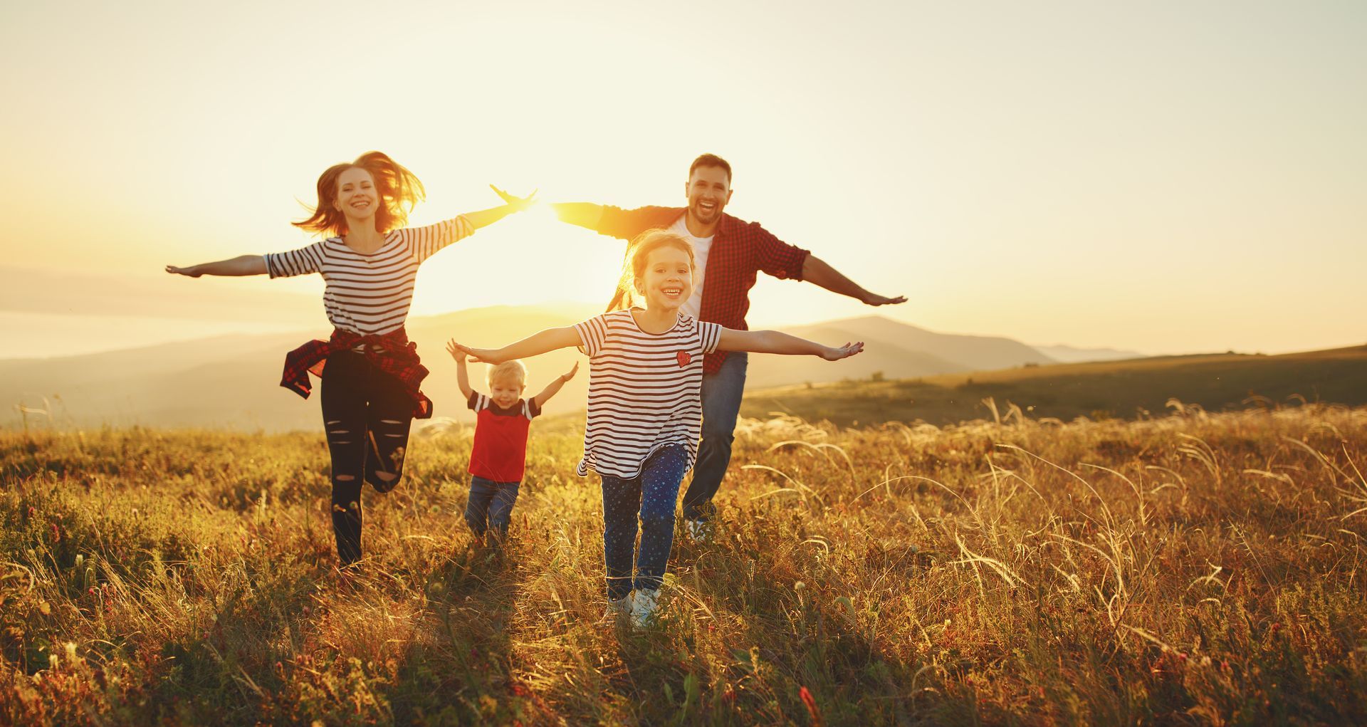 Family running with arms outstretched through a sunlit field at sunset
