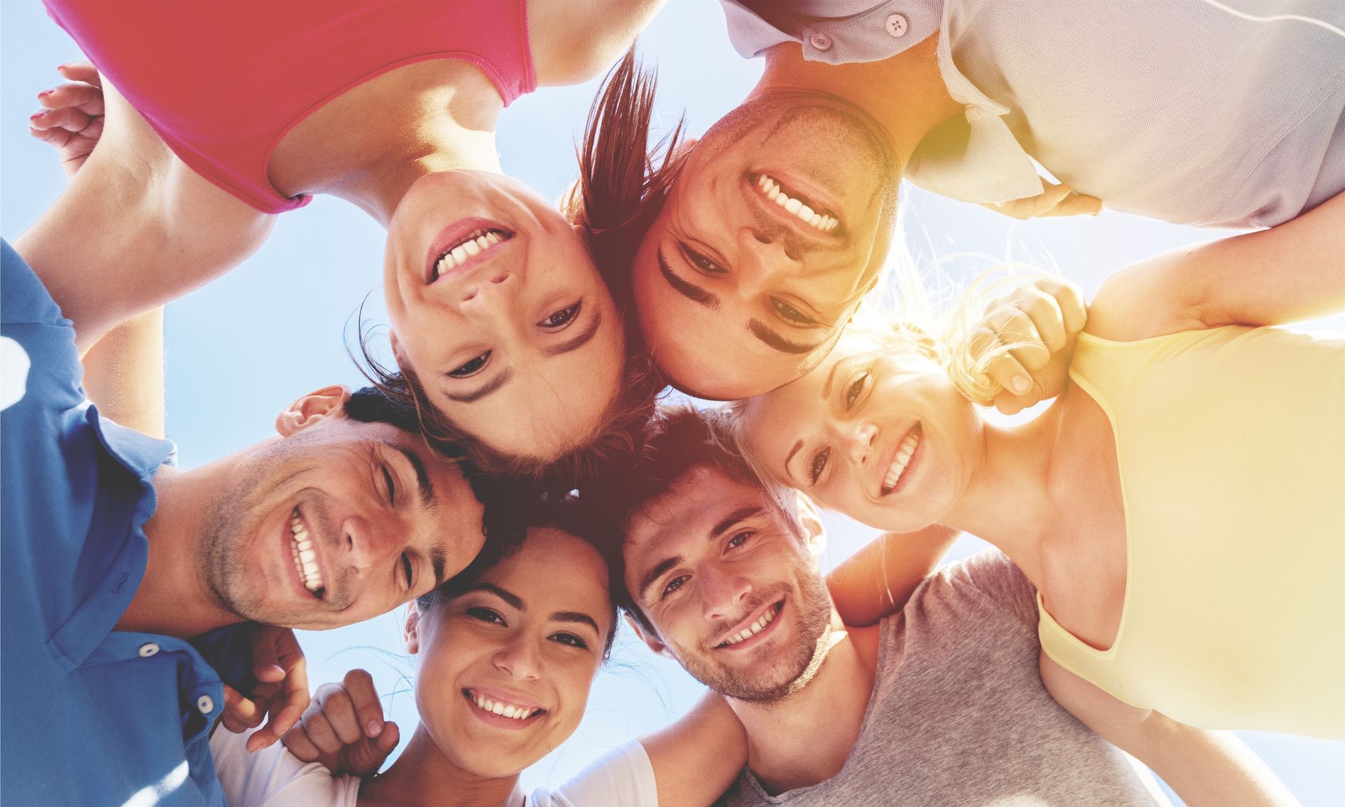Smiling friends lying in a circle outdoors, faces close together in bright sunlight