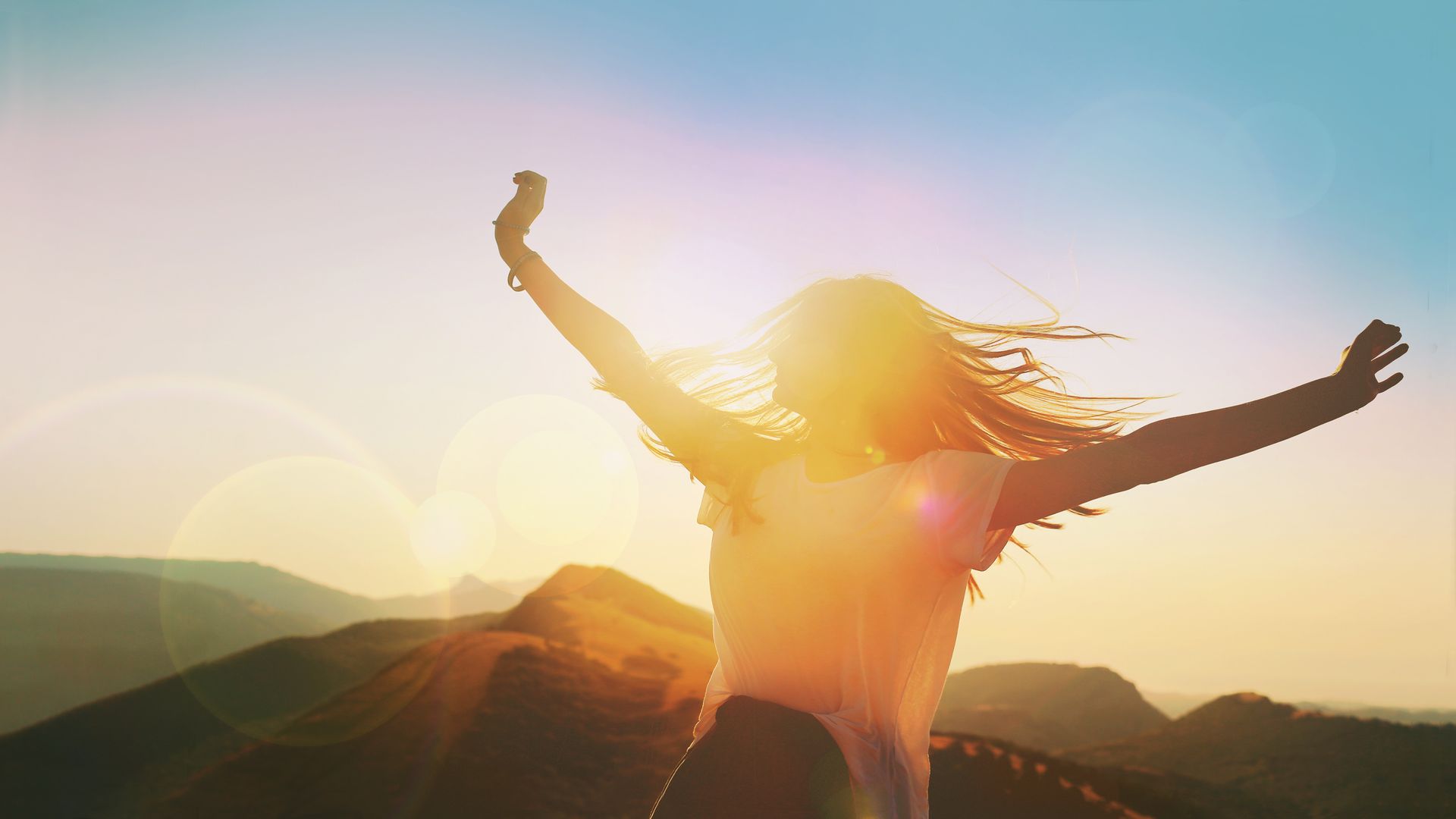 Silhouetted person with arms raised on a mountain at sunrise, with bright sun flare and blue sky.