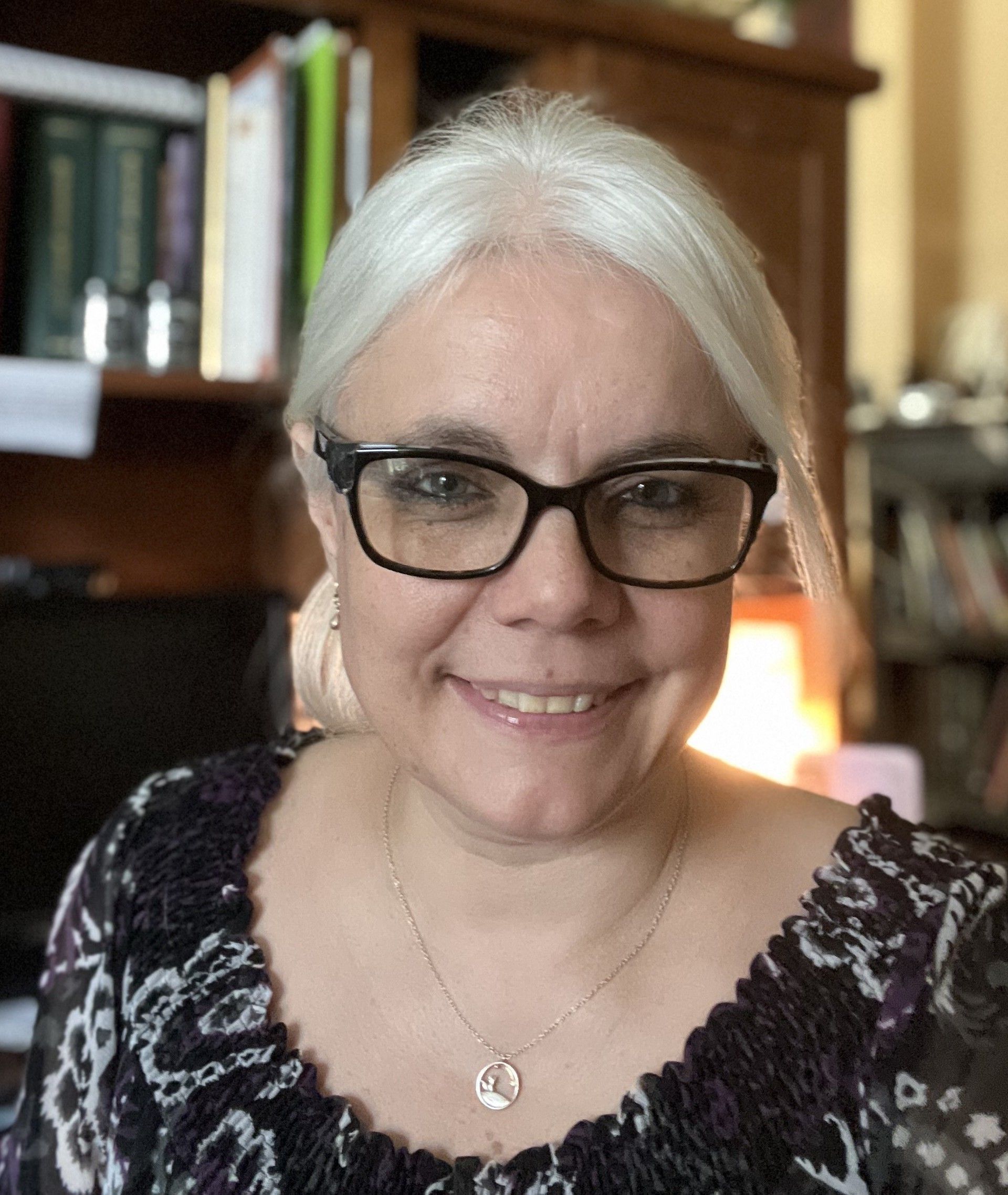 Smiling person with white hair and glasses, seated indoors with bookshelves in the background.