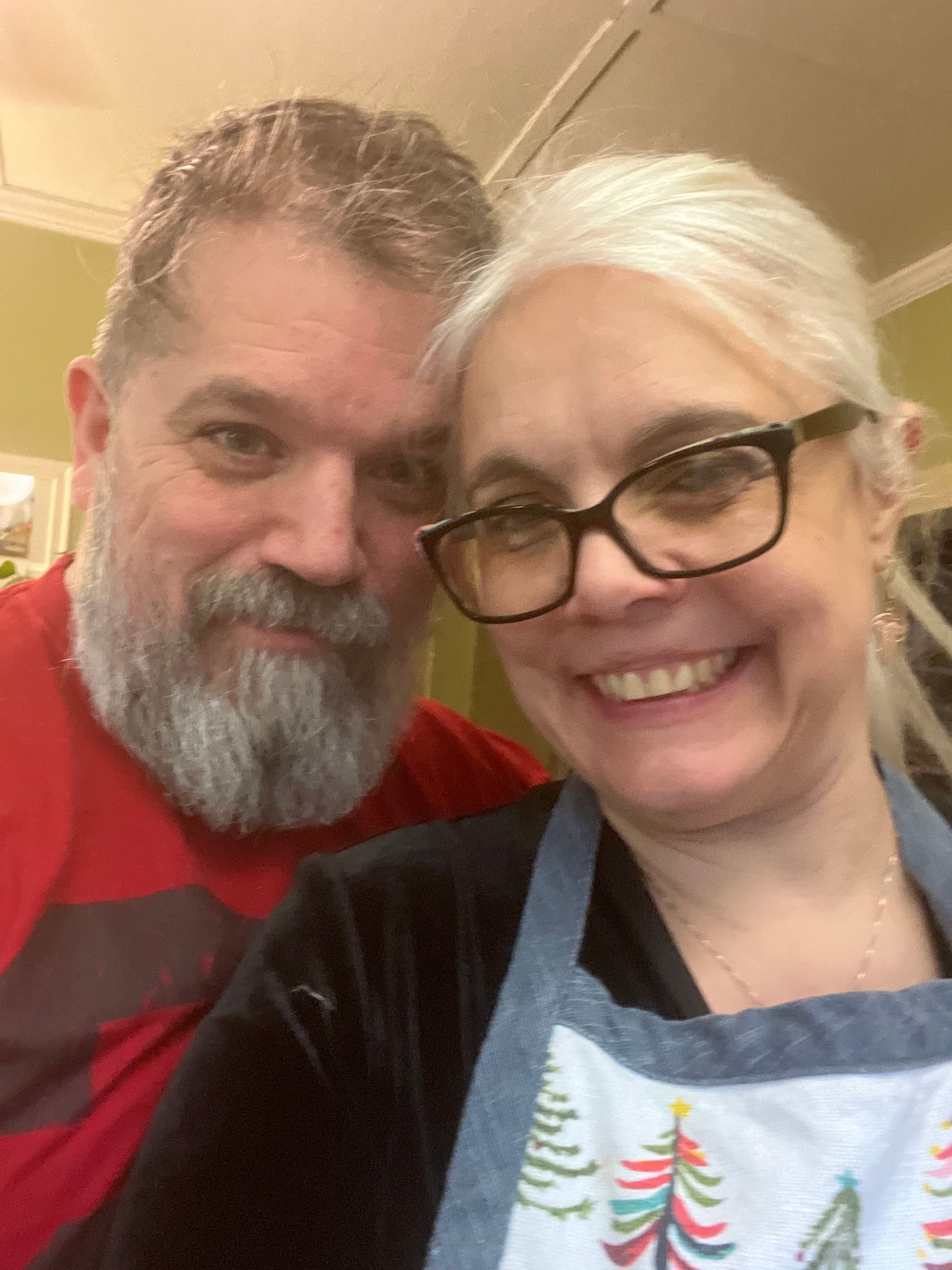 Two smiling people pose closely indoors, one in glasses and an apron, against a warm background.