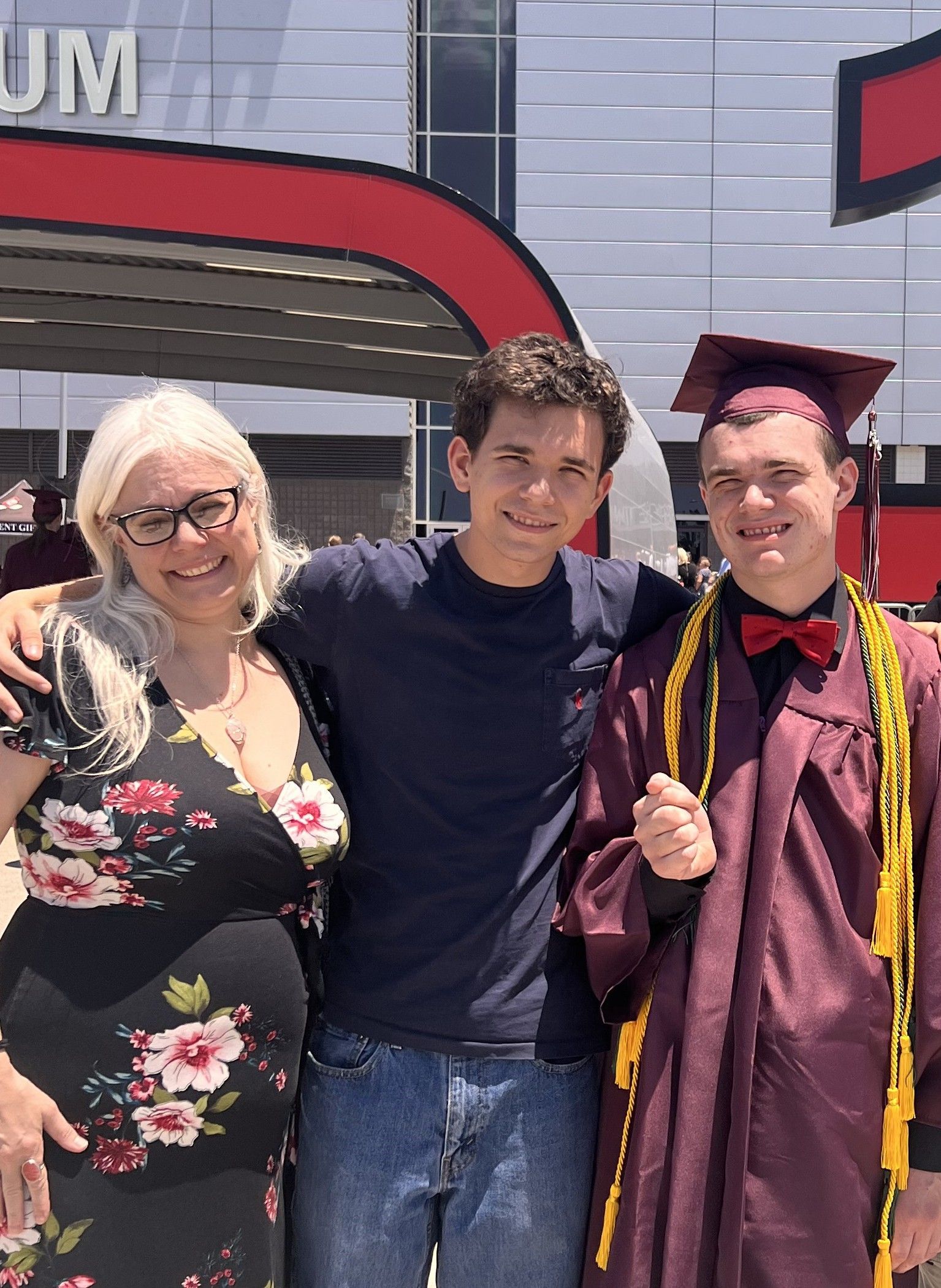 Three people posing outdoors in front of a building; one wears a graduation cap and maroon gown.