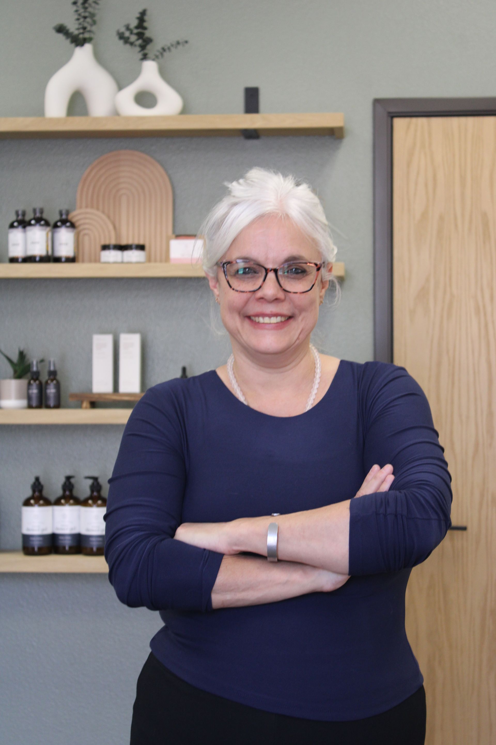 Smiling person with crossed arms in a navy top, standing in a modern shelf-lined room.