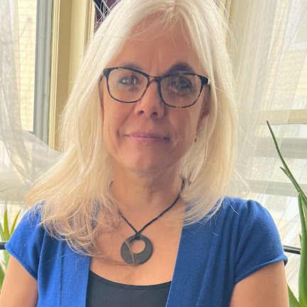 Blonde woman wearing glasses and a blue top, standing indoors by a bright window.