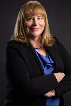 Woman in black blazer, blue blouse, arms crossed, smiling, against a dark gray background.