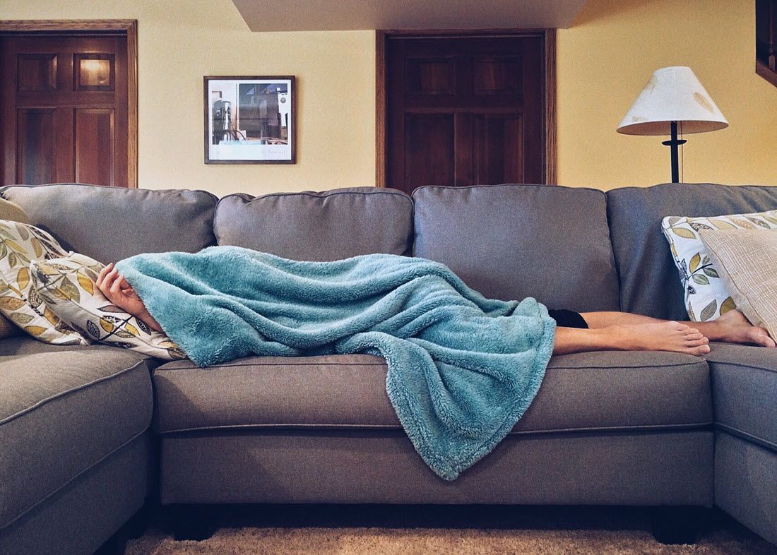 Person lying on a gray sectional sofa under a blue blanket in a living room.