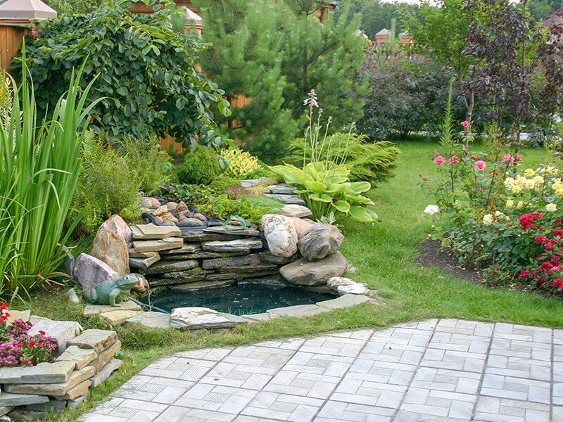 Backyard with a small pond and waterfall, surrounded by greenery, flowers, and a stone patio.