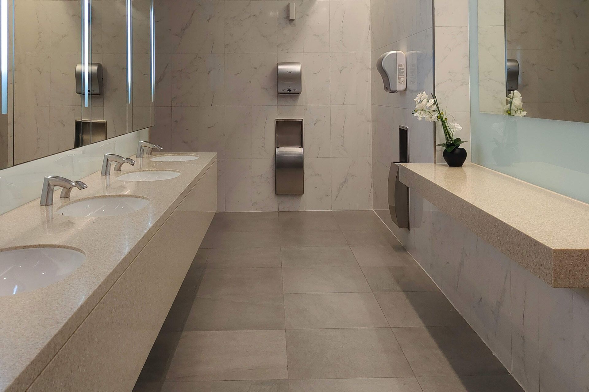 Modern public restroom with sinks, mirrors, and hand dryers. Light gray and beige tones.