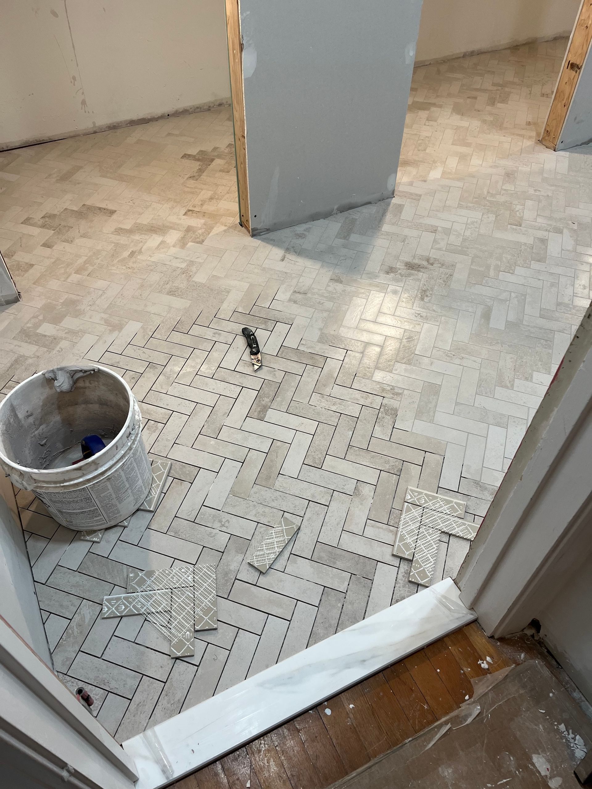 Floor tile installation in progress: Herringbone pattern, bucket, scattered tiles, doorways, gray and white.