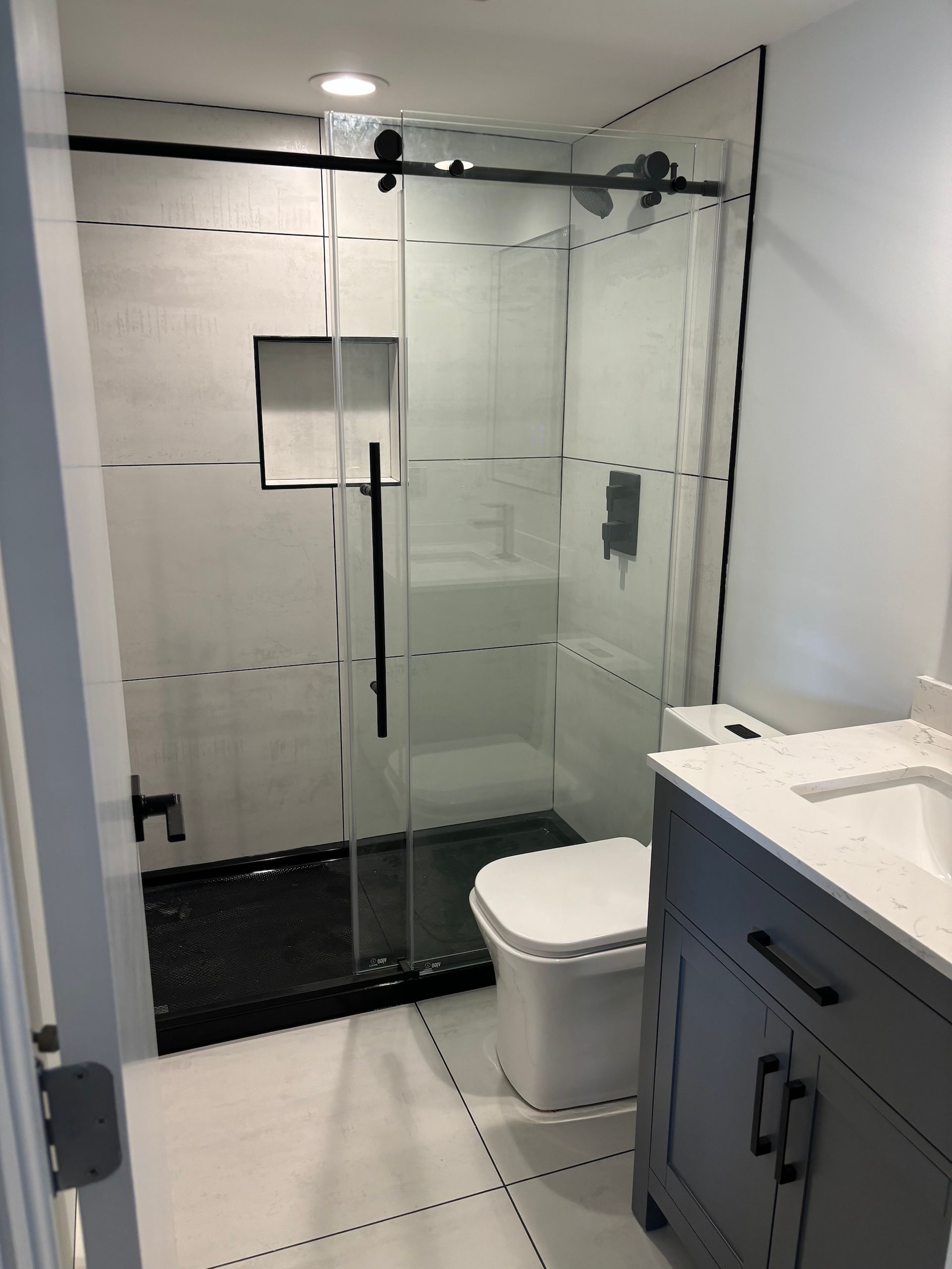 Modern bathroom with glass shower, black accents, white walls, and a gray vanity.