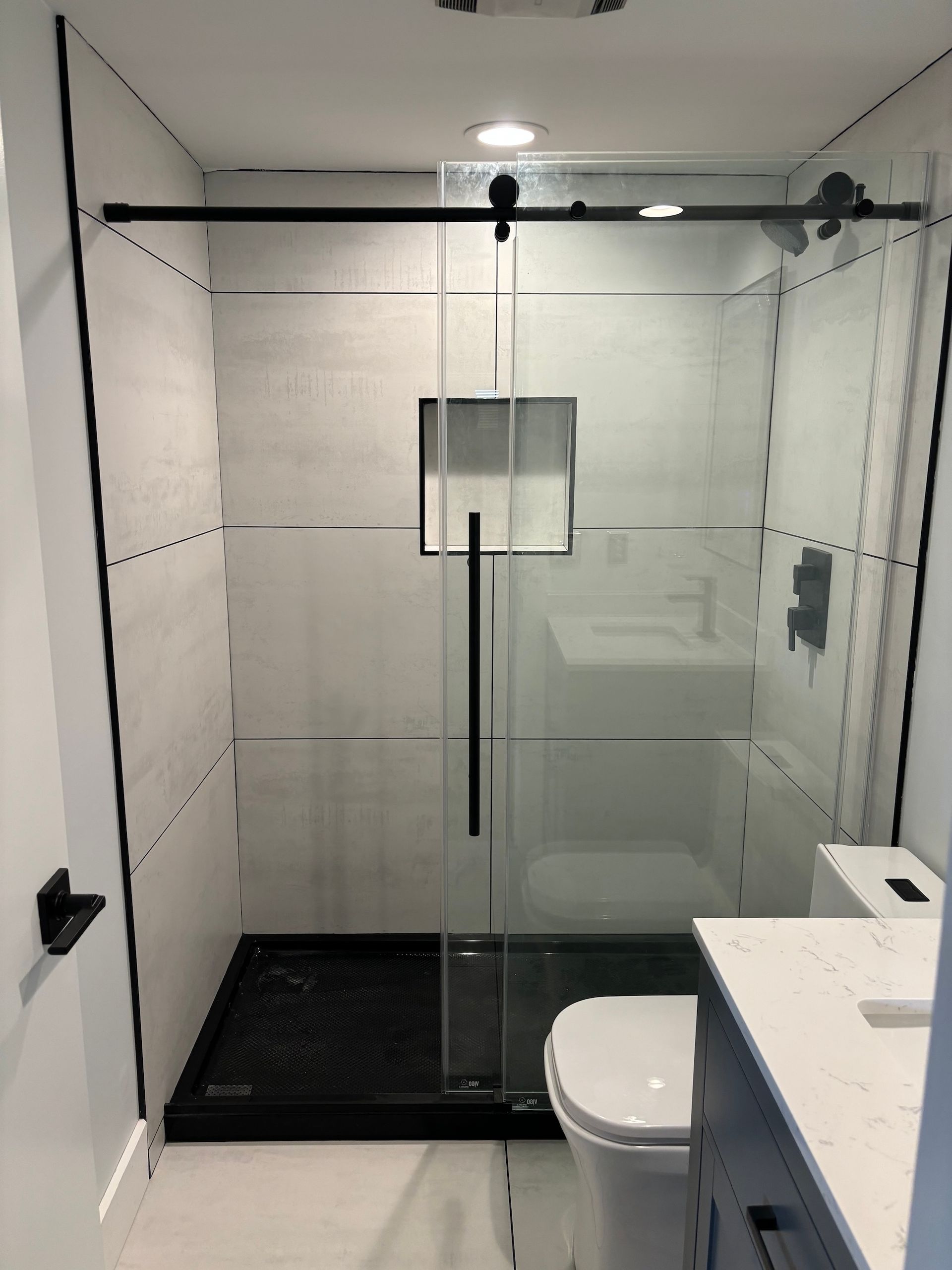 Modern glass walk-in shower with black tile floor, white walls, and a toilet beside a white sink.