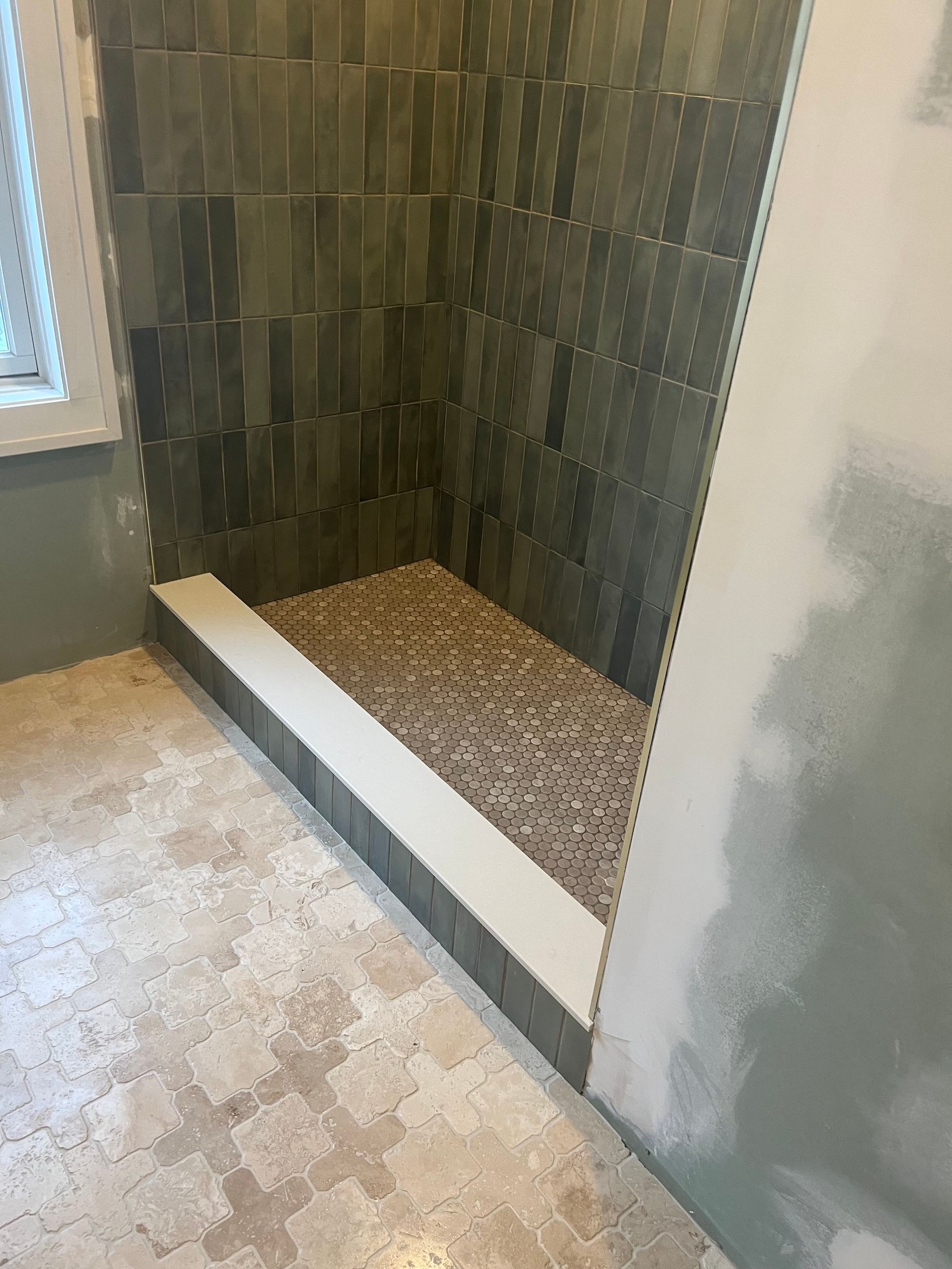 Partially tiled shower corner with pebble shower floor and unfinished drywall beside a window