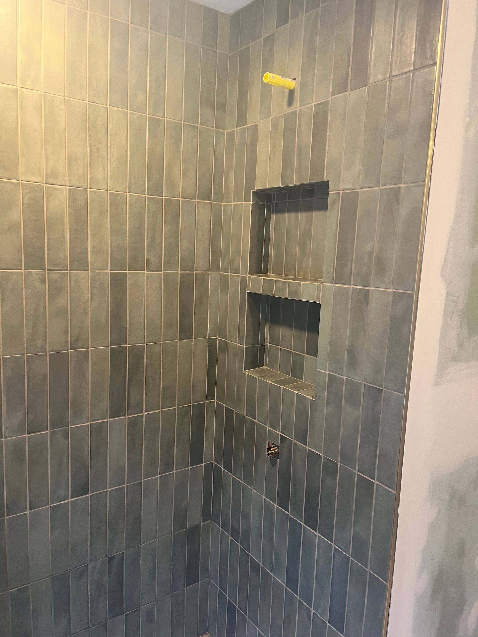 Gray tiled shower with two built-in shelves. Yellow showerhead. Unfinished wall on the right.