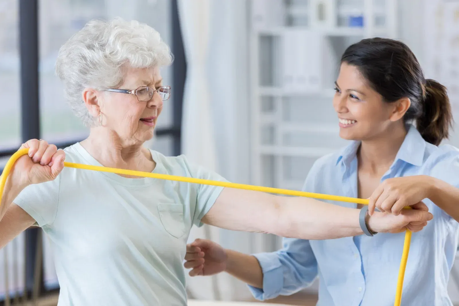 Caregiver assisting an older person to stand in a living room; caregiver wears blue scrubs and smiles.