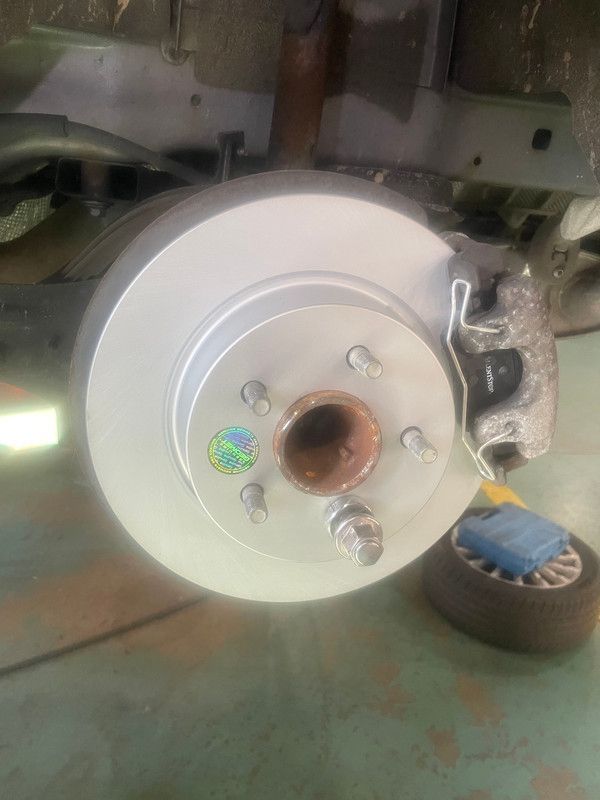 A car's brake rotor and caliper; new silver rotor, mounted, with a tire resting nearby.