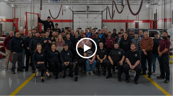 Group of people in a garage; posing for a photo. Some standing, some sitting. Many are wearing dark shirts.