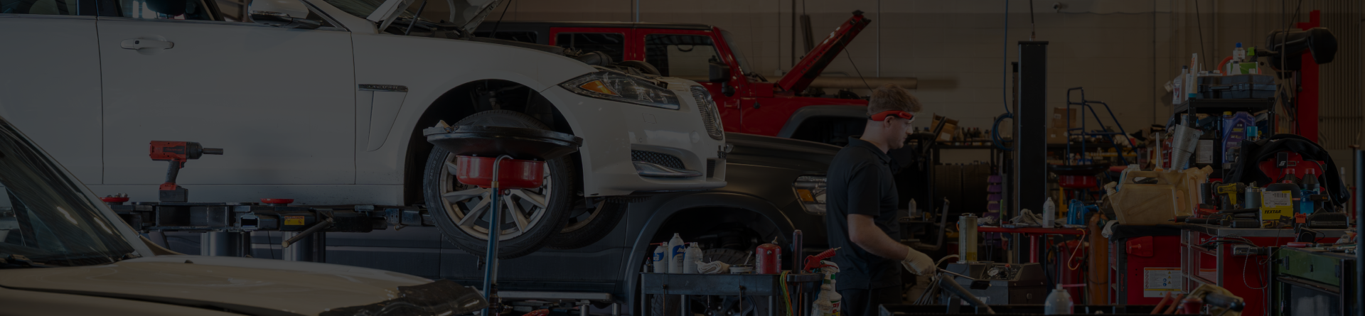 Mechanic working in a garage, white car on a lift, red SUV in the background, and tools are visible.
