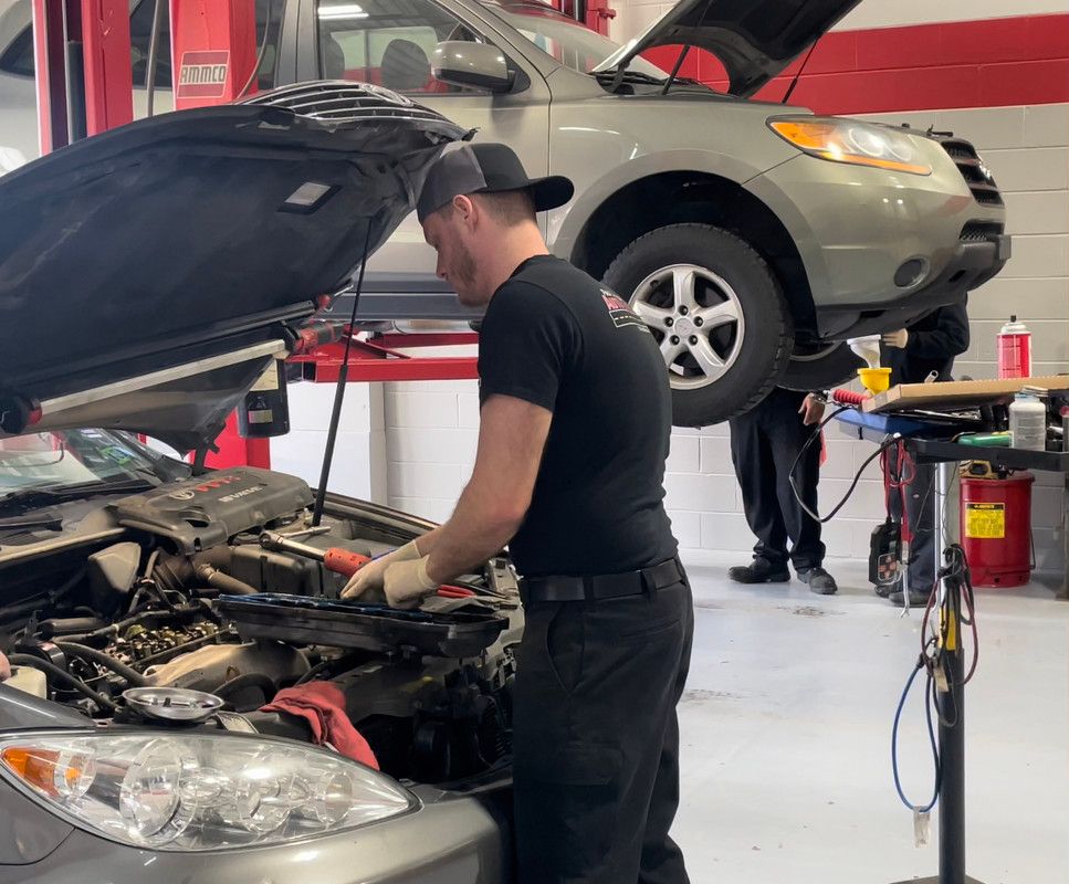 Mechanic working on a car engine in a garage, with another car raised on a lift.