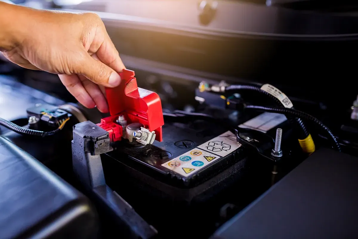 Hand removing a red protective cap from a car battery terminal in an engine bay.