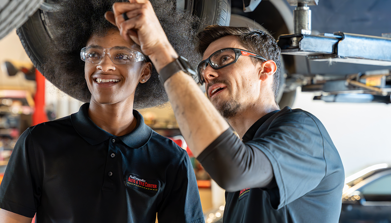 Two mechanics working under a car; one points, both wear safety glasses and smile.