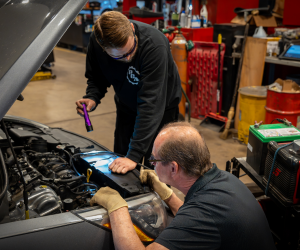 Two mechanics inspecting a car engine bay. One holds a flashlight, another works with tools.