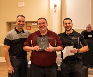 Three men in black and maroon shirts holding a plaque at an event. Two smile; one holds a microphone.