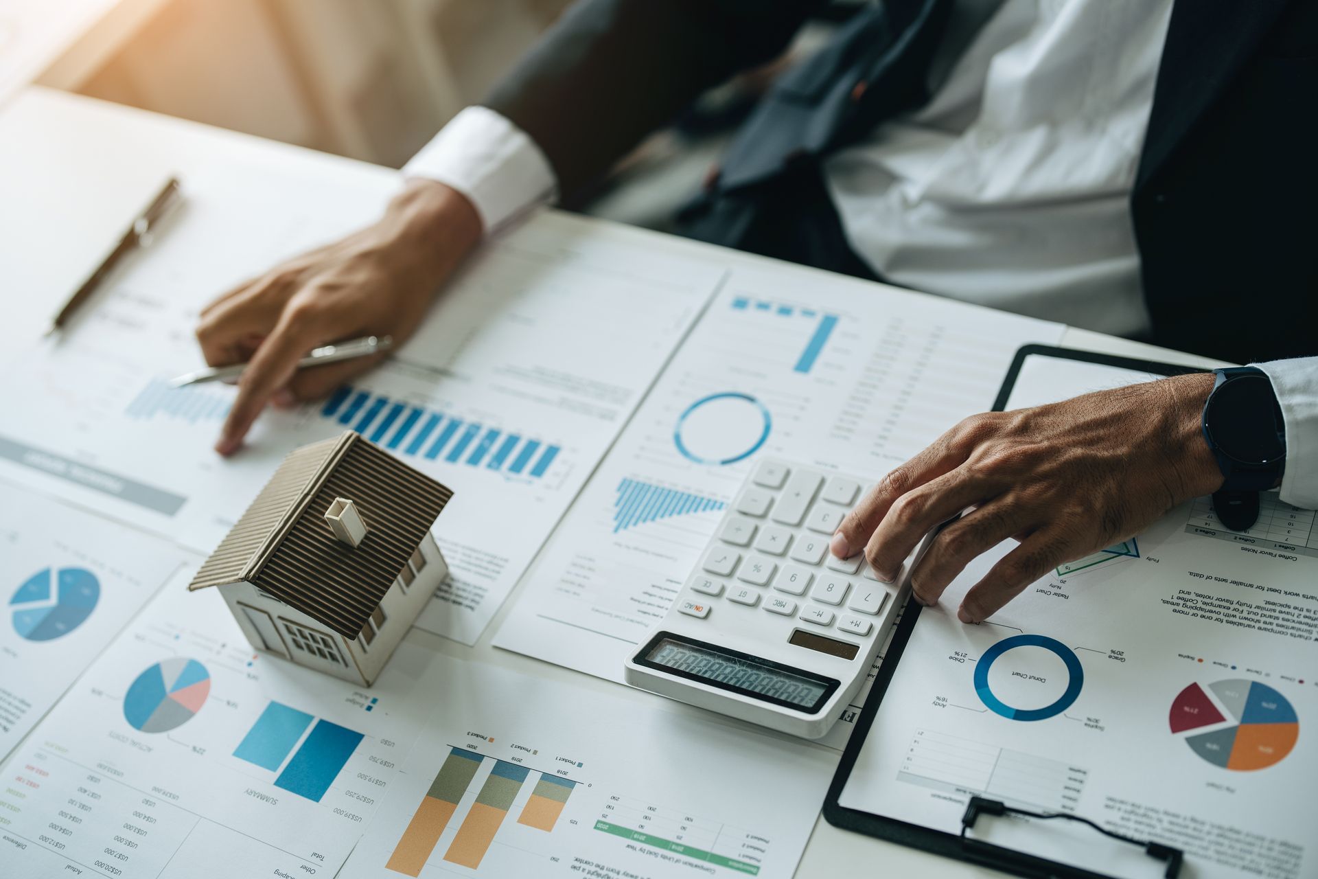 Hands pointing at charts and using a calculator, next to a miniature house, on a desk with papers.
