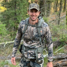 A man in a camo shirt and hat is standing in the woods with a backpack.