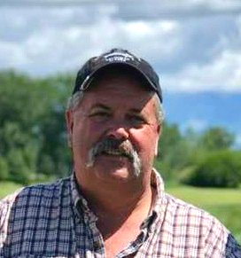 A man with a mustache is wearing a plaid shirt and a baseball cap.