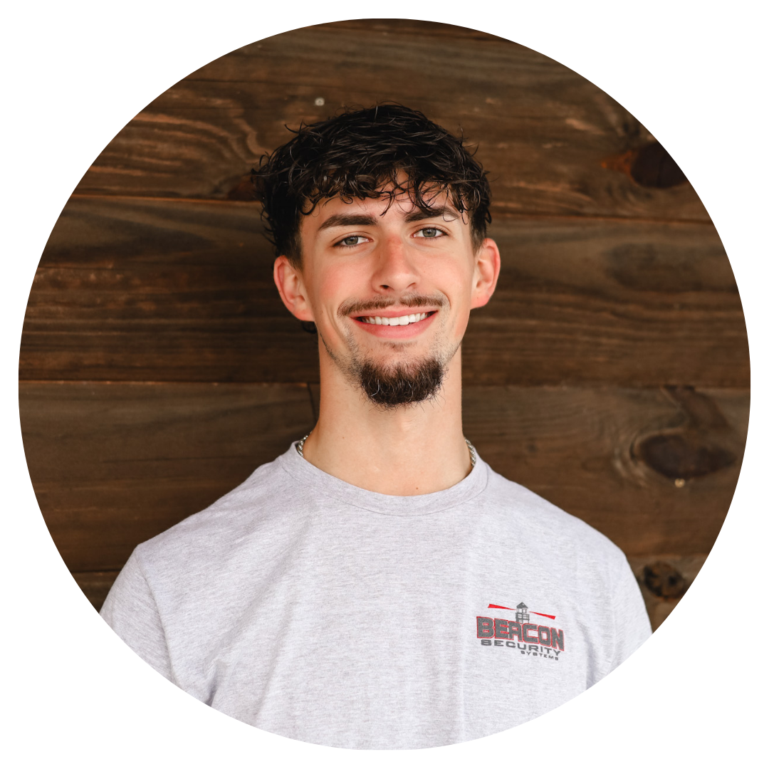 A young man with a beard is smiling in front of a wooden wall.