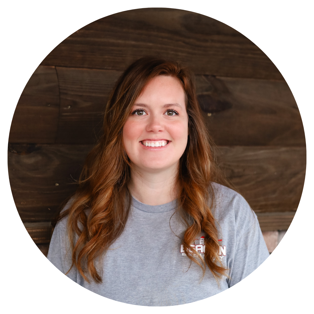 A woman in a grey shirt is smiling in front of a wooden wall.