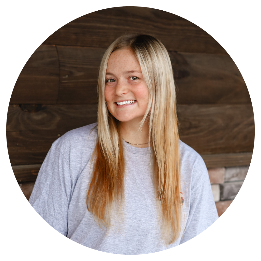 A woman with long blonde hair is smiling in front of a wooden wall