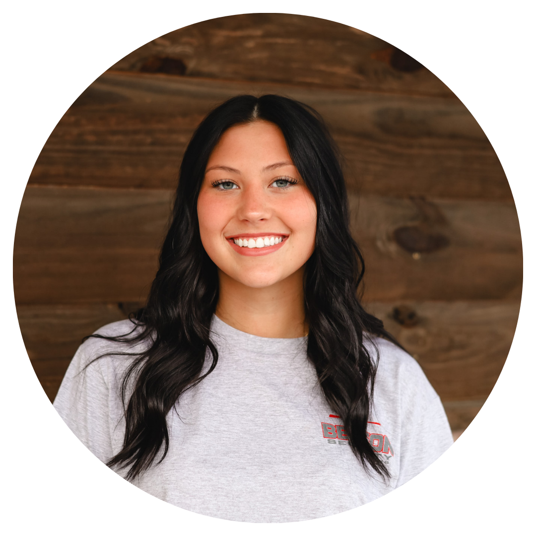 A woman in a grey shirt is smiling in front of a wooden wall