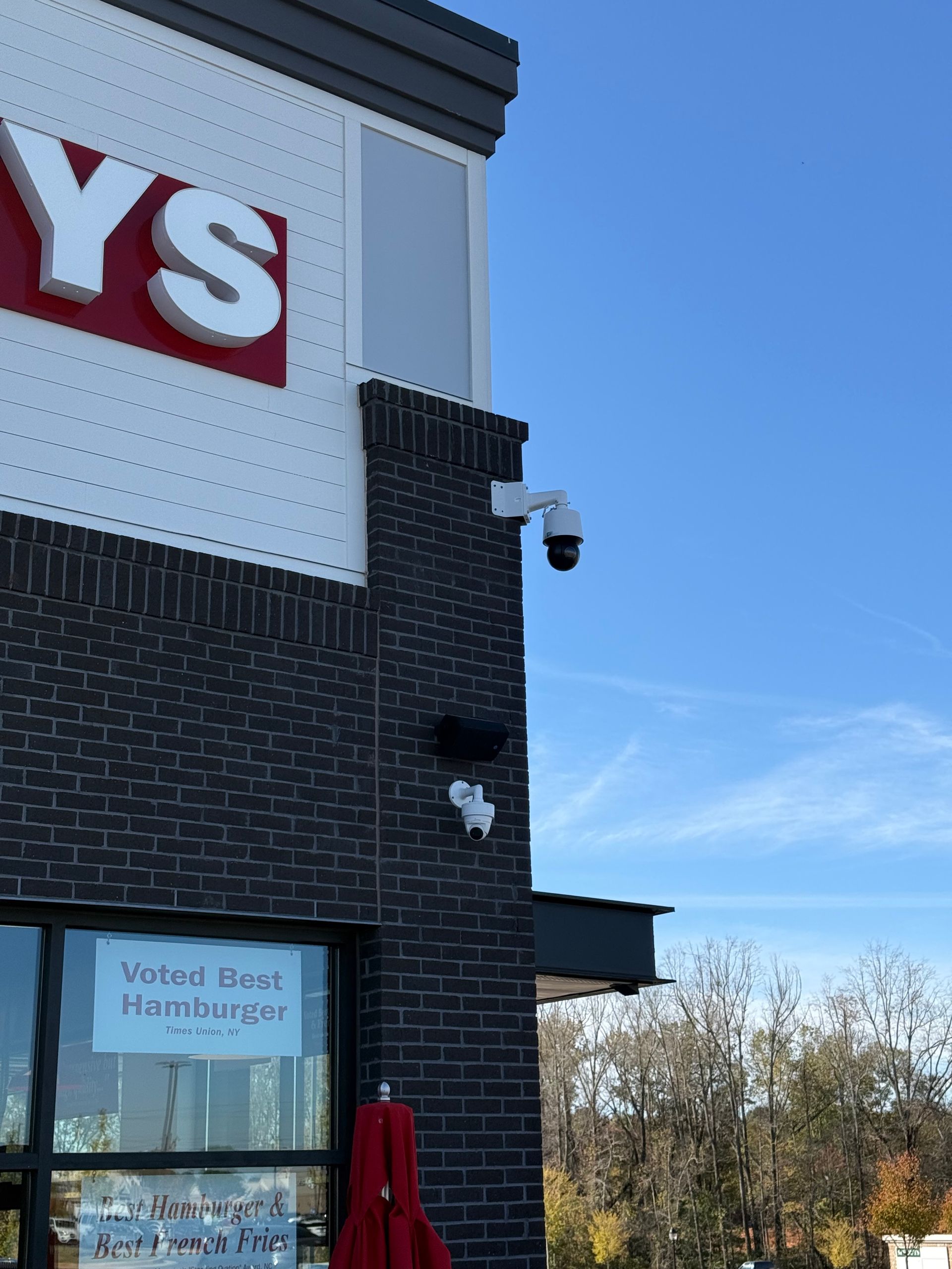 Exterior of a restaurant with a partial “YS” sign, blue sky, security cameras, and a “Voted Best Hamburger” window sign