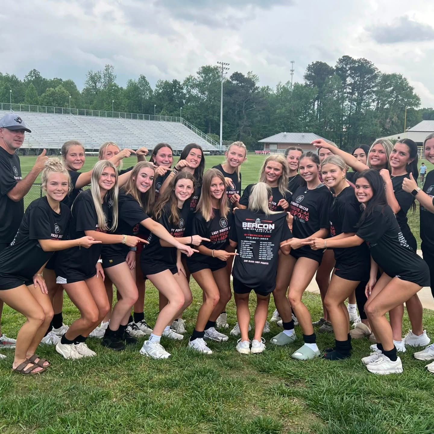 Group of athletes in black uniforms posing on a grassy field, smiling and holding a championship sign.