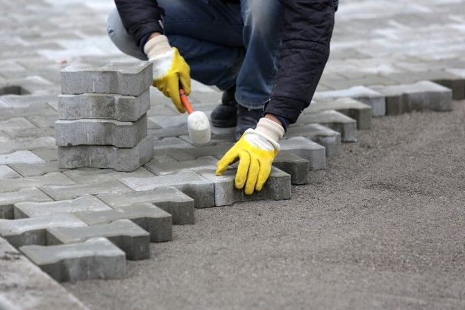 Worker laying gray paving stones with yellow gloves and a mallet on a sidewalk construction site