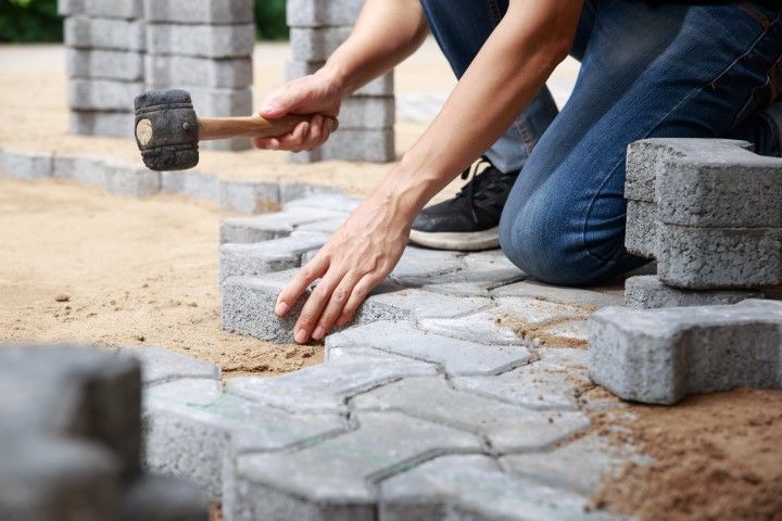 Person laying gray paving stones with a mallet and sand at an outdoor worksite