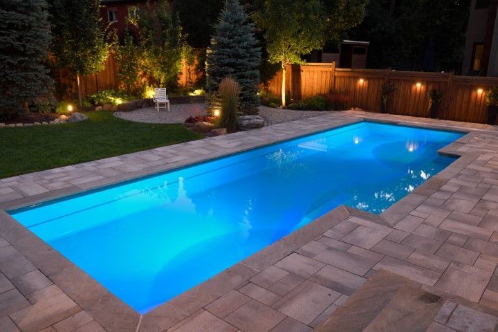 Backyard swimming pool glowing blue at dusk, bordered by stone pavers and landscaped garden lighting