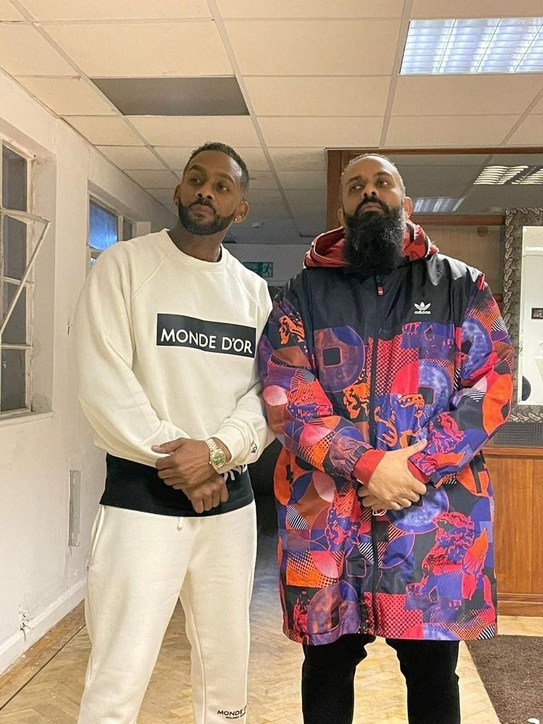Two men standing in a hallway. One in cream-colored outfit, the other in a patterned jacket and beard.