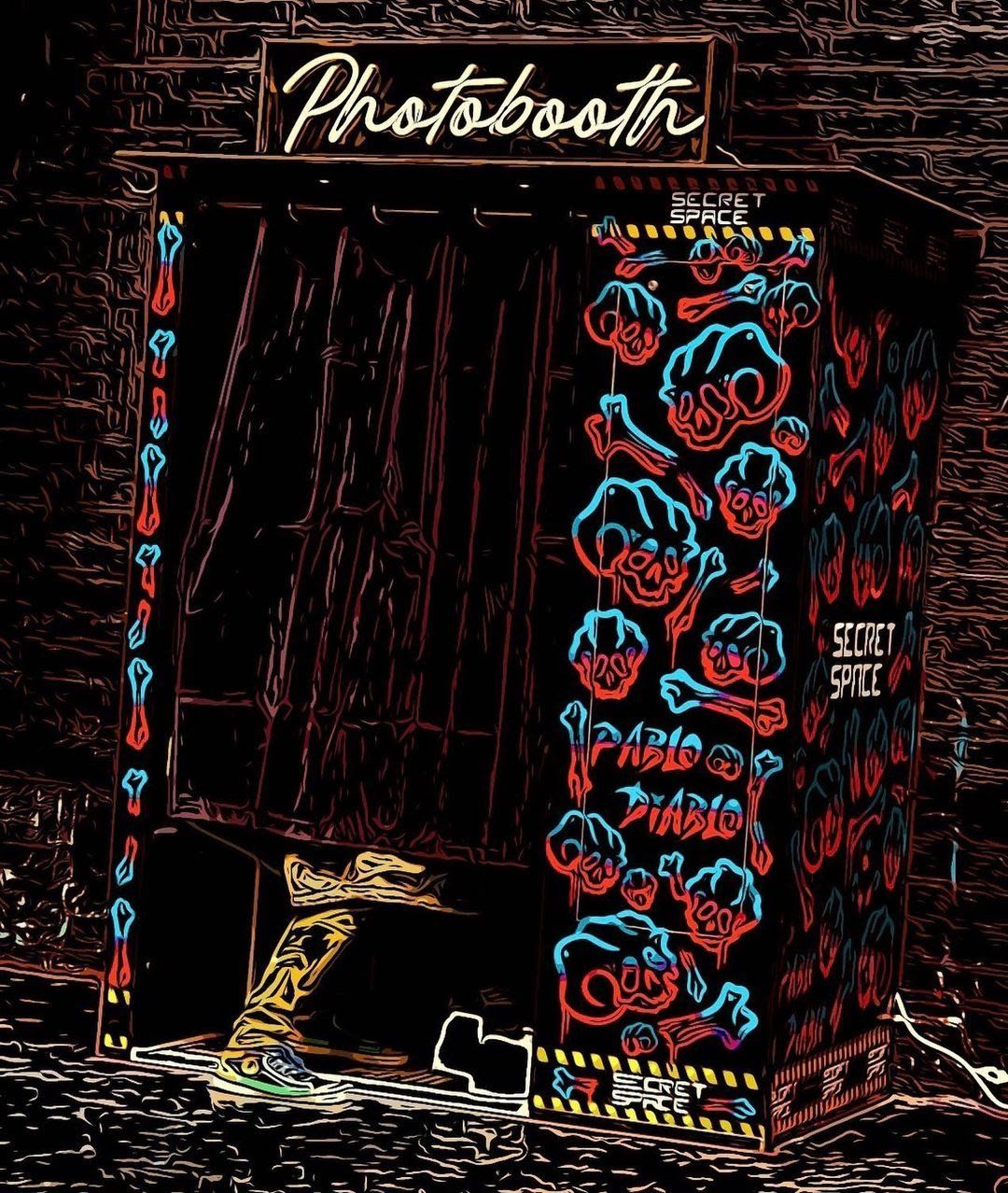Photobooth, neon sign above. Exterior with red & blue art. Dark curtains open, lower leg visible.