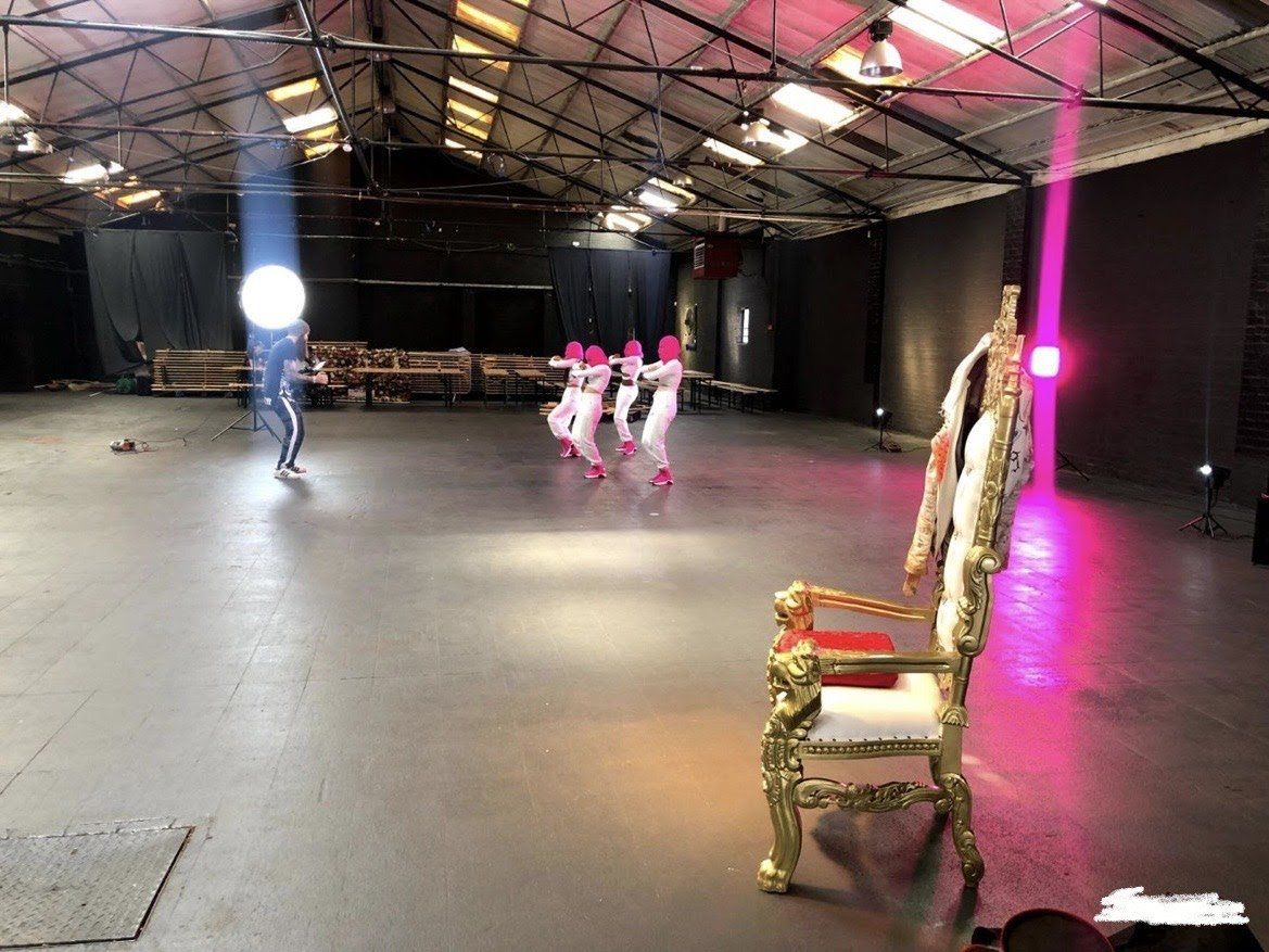 Dancers in white with pink hats perform on a stage. A gold chair sits in front.