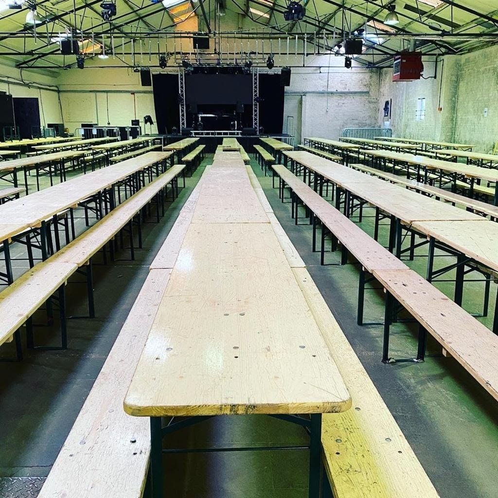 Long wooden tables and benches arranged in rows inside a large, industrial-style venue, leading to a stage.