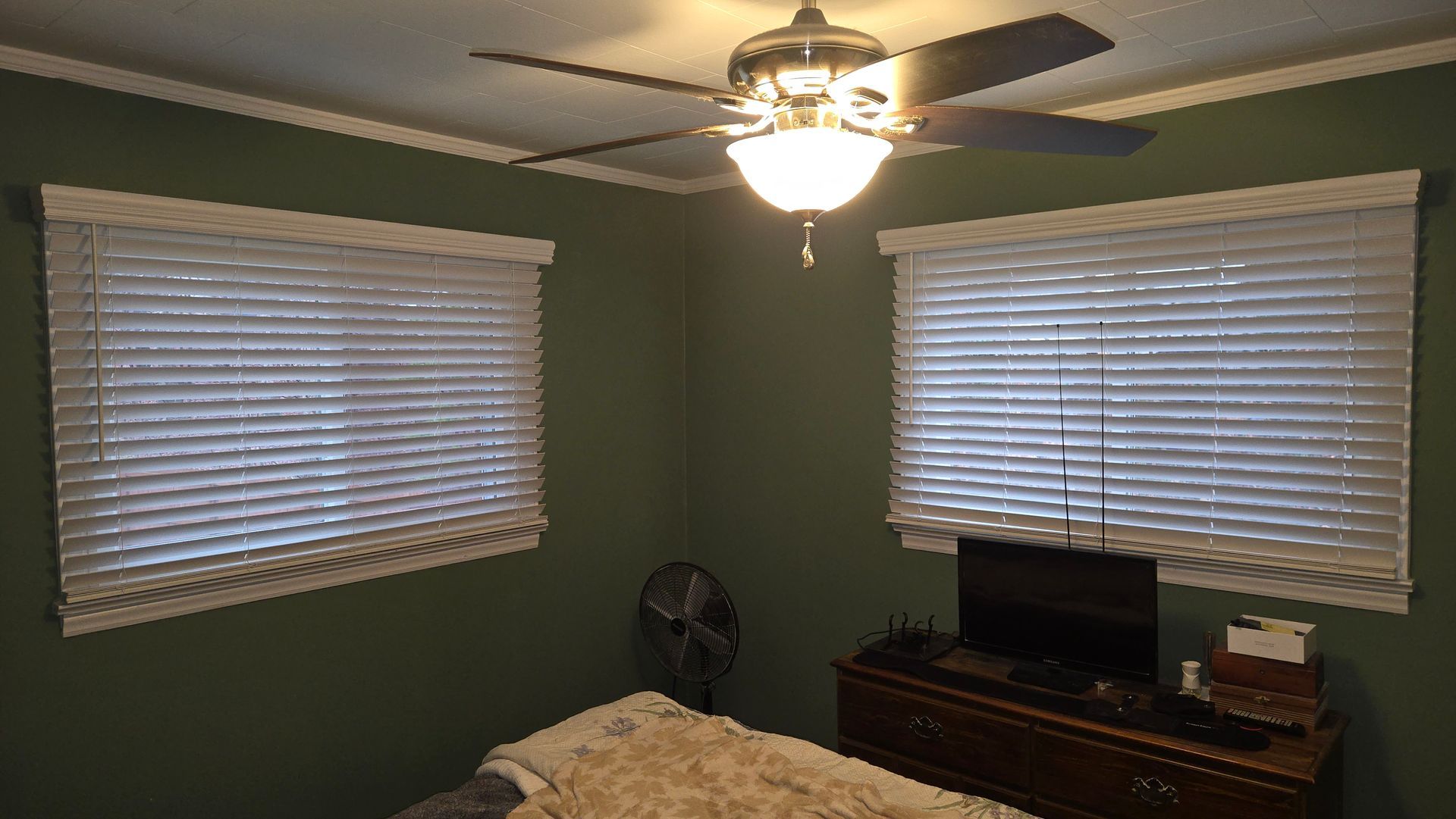 Bedroom with green walls, two windows with blinds, a ceiling fan, and a dresser with a TV.