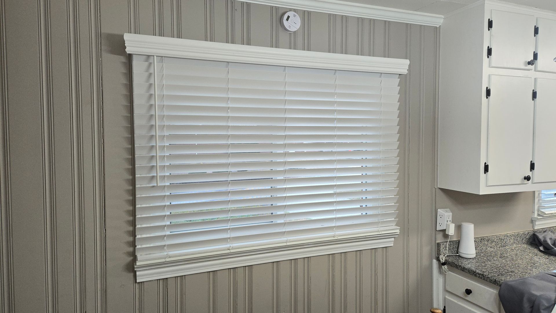 White blinds on a window in a room with gray striped wallpaper and white cabinets nearby